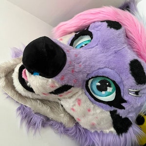 May include: Purple and pink furry animal head with black nose and blue eyes. The head is tilted to the side and the eyes are looking to the right.