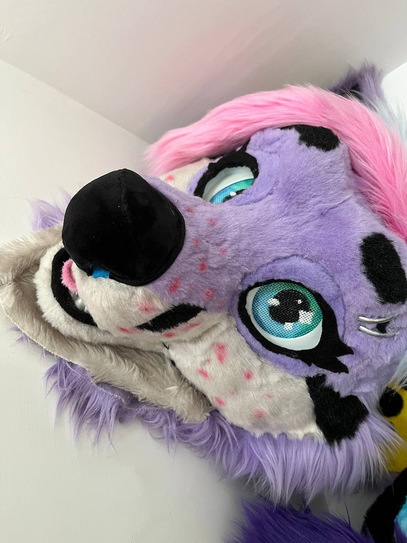 May include: Purple and pink furry animal head with black nose and blue eyes. The head is tilted to the side and the eyes are looking to the right.