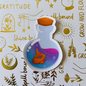 May include: A sticker of a glass bottle with a cork stopper, filled with a purple liquid and a small orange goldfish. The sticker is on a white background with gold foil designs, including stars, flowers, and the words "spellbound", "gratitude", "shine bright", "magical", and "grow and flourish".