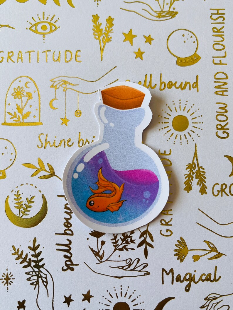 May include: A sticker of a glass bottle with a cork stopper, filled with a purple liquid and a small orange goldfish. The sticker is on a white background with gold foil designs, including stars, flowers, and the words "spellbound", "gratitude", "shine bright", "magical", and "grow and flourish".