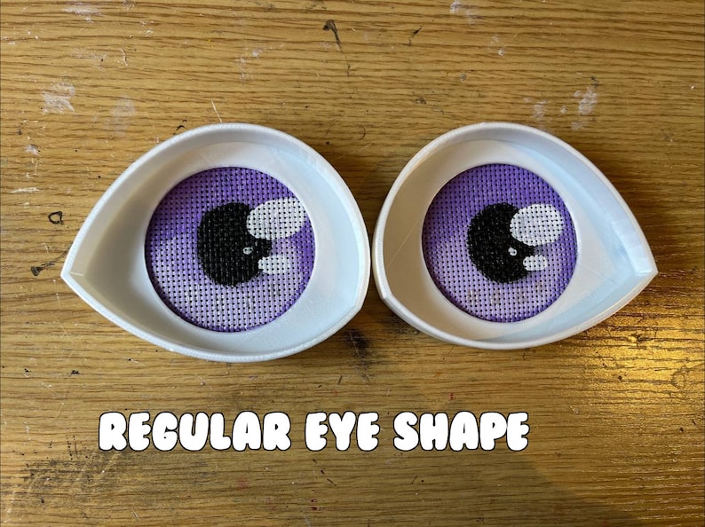 May include: Two white plastic eye shapes with purple fabric eyes inside. The eyes have black pupils and white highlights. The text "REGULAR EYE SHAPE" is below the eyes.