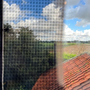 May include: A view through a window with a screen, showing a red tiled roof, a green field, and a blue sky with white clouds.