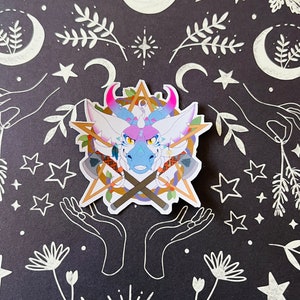 May include: A sticker featuring a cartoon dragon with pink and blue scales, a gold star, and crossed axes. The dragon has a white background and is surrounded by a wreath of leaves. The sticker is on a black background with white stars, moons, and other designs.