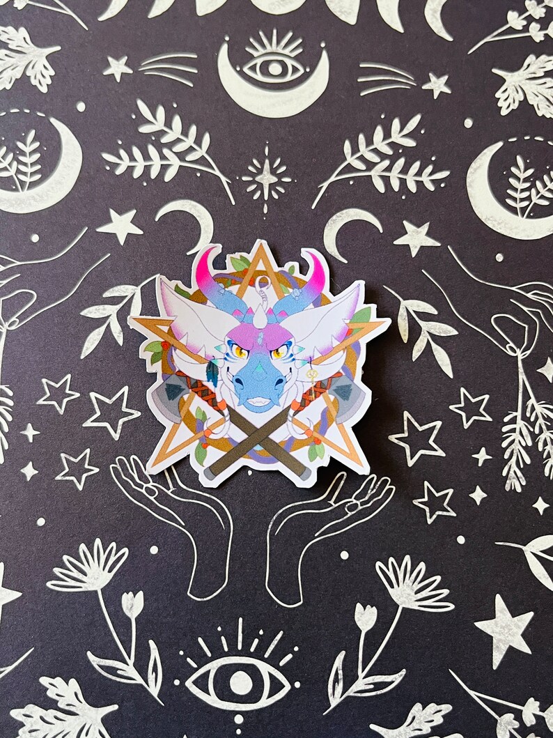 May include: A sticker featuring a cartoon dragon with pink and blue scales, a gold star, and crossed axes. The dragon has a white background and is surrounded by a wreath of leaves. The sticker is on a black background with white stars, moons, and other designs.