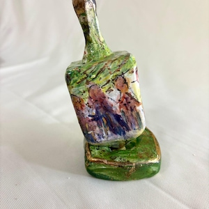 May include: A hand-painted, decorative bottle stopper. The stopper features a green base and handle with gold accents. The main body of the stopper is painted with a scene of figures in a landscape, with shades of purple, blue, and yellow.
