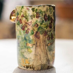Peut inclure: Une tasse en céramique avec un motif peint à la main d'un arbre aux feuilles vertes et aux fleurs roses. La tasse a une anse dorée et les mots "Get out Trouble" écrits en lettres dorées au fond.