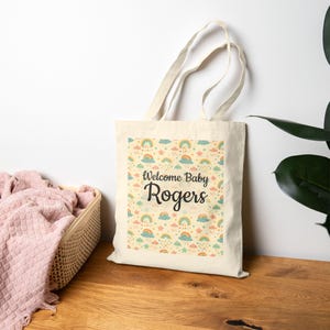 May include: A cream-colored tote bag with a colorful pattern of rainbows, clouds, and stars. The text "Welcome Baby Rogers" is printed in black script. The bag has long handles and is made of a sturdy fabric.