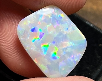 May include: A polished, rectangular opal gemstone with a white base and iridescent flashes of color, including blue, green, and yellow. The gemstone is held between fingers, showcasing its vibrant play-of-color and unique pattern.
