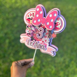 Pode incluir: Topper de bolo temático Minnie Mouse rosa e branco com um laço, orelhas e o número 3. O topper também inclui o nome "Le'Ana" em letras rosa e brancas.