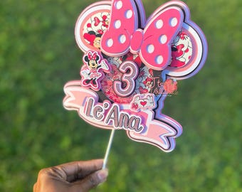 May include: Pink and white Minnie Mouse themed cake topper with a bow, ears, and the number 3. The topper also includes the name "Le'Ana" in pink and white lettering.