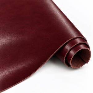 Puede incluir: Un rollo de cuero color burdeos intenso con una textura de superficie lisa y mate. El material está enrollado firmemente en un extremo, revelando el lado inferior natural y fibroso de la piel, mientras que la capa exterior muestra un color rico y uniforme.
