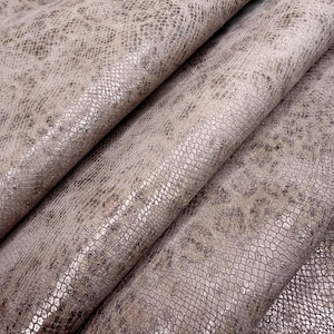 May include: A piece of fabric featuring a metallic beige and bronze snakeskin pattern. The material has a textured surface with a repeating scale design that creates a shimmering effect across the folds of the draped textile material.