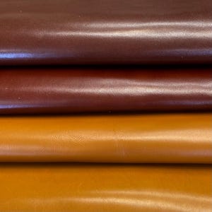 May include: Close-up of three leather samples in varying shades. The top is a dark burgundy, the middle a mid-brown, and the bottom a warm, golden-yellow. The leather has a smooth texture and a subtle sheen, indicating a quality finish.
