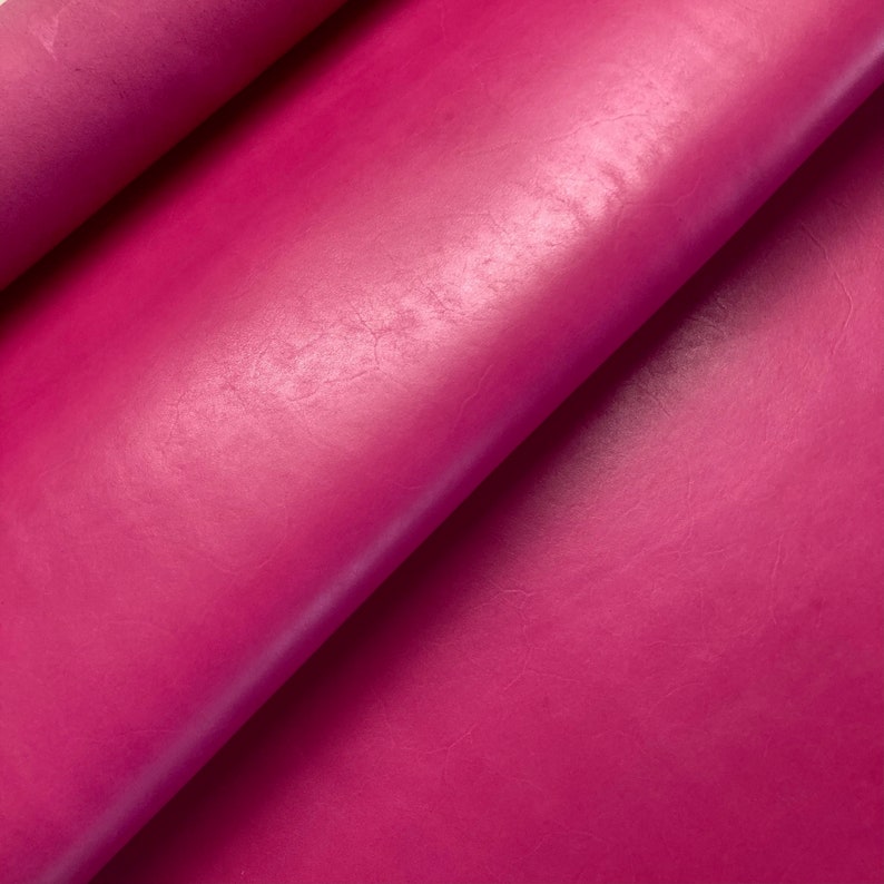 May include: Close-up of a vibrant pink leather fabric with a smooth, slightly textured surface. The fabric is folded over, showcasing its soft and supple texture.