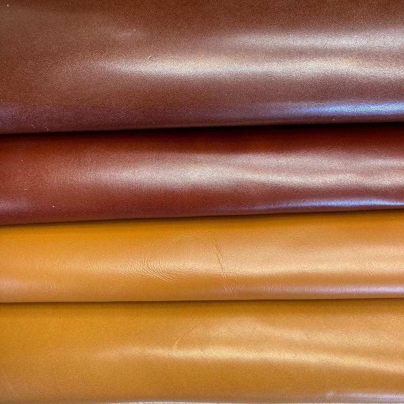 May include: Close-up of four different shades of leather. The colours range from a deep burgundy to a light tan, with variations of brown and orange. The leather appears smooth and has a slight sheen.