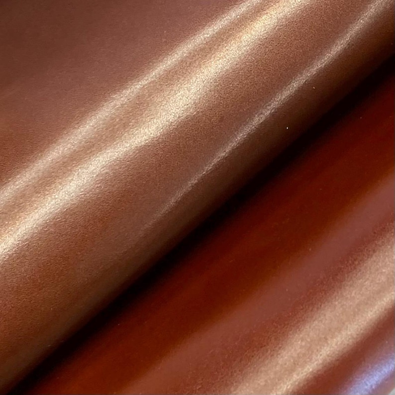 May include: Close-up of a sheet of brown leather with a glossy finish. The leather has a smooth texture and a slight sheen, with folds and creases visible across the surface. The colour is a rich, warm brown with highlights.