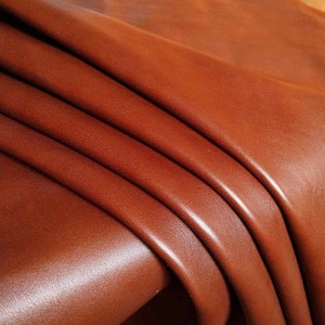 Könnte beinhalten: Nahaufnahme eines gefalteten Stücks braunen Leders. Das Leder hat eine glatte, glänzende Oberfläche und eine leicht strukturierte Maserung.