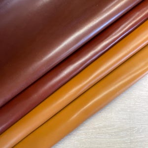 May include: Close-up of three sheets of leather in varying shades of brown. The top sheet is a deep burgundy, followed by a medium reddish-brown, and then a lighter, golden-brown hue. The leather appears smooth and glossy.