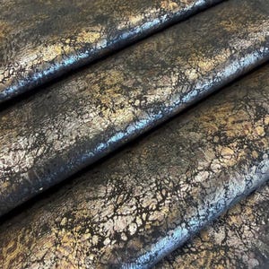 May include: A folded piece of fabric featuring a dark black base colour with a metallic gold and bronze crackled texture overlay. The material displays a distressed, weathered appearance with deep crevices and raised, shimmering highlights.