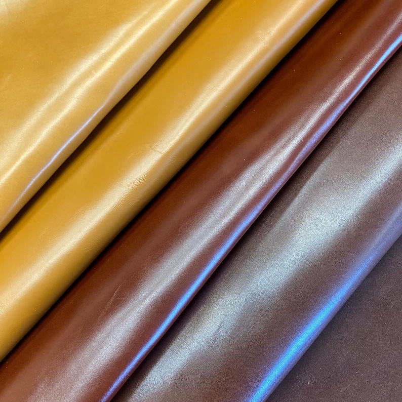 May include: Close-up of four sheets of leather in shades of yellow, gold, brown, and purple. The leather has a smooth, shiny surface, with subtle variations in colour and texture. The sheets are arranged diagonally, creating a visually appealing pattern.