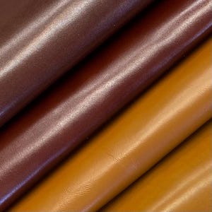 Puede incluir: Primer plano de tres muestras de cuero en tonos marrón y beige. El cuero tiene una superficie lisa y brillante, con pliegues y texturas visibles. Los colores van desde un marrón intenso y rico hasta un beige dorado más claro.