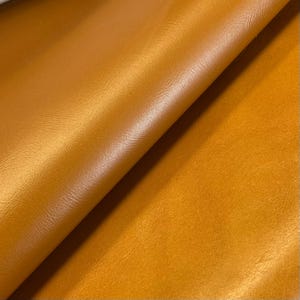 May include: Close-up of a sheet of smooth, golden-brown leather. The material has a slight sheen, with subtle variations in colour and texture. The leather is folded, creating shadows and highlights that emphasise its supple quality.