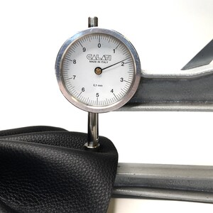 May include: A close-up of a leather sample being tested with a dial gauge. The gauge reads 0.1 mm and is labeled "CALATI MADE IN ITALY".