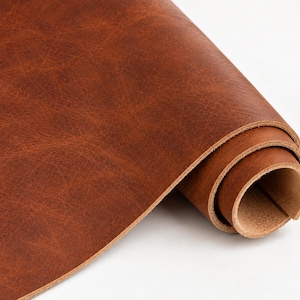 May include: A roll of medium brown leather featuring a visible grain pattern and a smooth, matte finish. The material is coiled at one end, revealing the lighter, textured underside of the hide, showcasing the natural thickness and flexible quality.