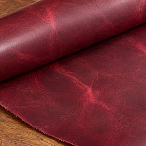 Può includere: Un rotolo di pelle color bordeaux intenso caratterizzato da una texture marmorizzata e vissuta, con venature rosso chiaro su tutta la superficie. Il materiale presenta una finitura liscia e opaca ed è parzialmente srotolato per mostrare la grana.