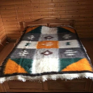 Puede incluir: Una manta de lana blanca, gris, verde y naranja con un borde con flecos. La manta está sobre un marco de cama de madera.