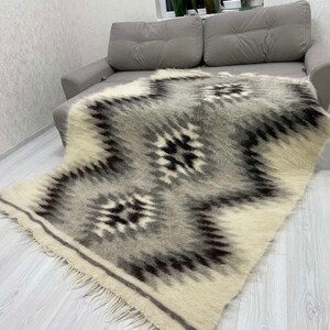 May include: A rectangular, woven blanket with a geometric pattern in shades of gray, black, and cream. The blanket has a fringed edge and is draped over a gray sofa. The design features repeating diamond and star-like shapes.