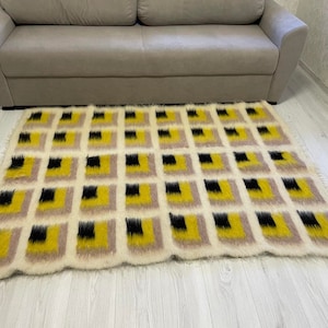 May include: A white, fluffy rug with a geometric pattern of black and yellow squares. The rug is laid out on a light wood floor.