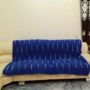 Può includere: Una coperta blu con un motivo bianco è drappeggiata su un divano color crema. Il divano si trova in un soggiorno con tende marroni e una pianta in vaso.