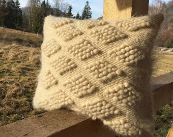 May include: A cream-colored woven pillow with a textured diamond pattern. The pillow is resting on a wooden fence against a backdrop of a grassy field and trees.