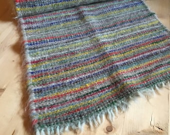 May include: A handwoven rug with a striped pattern in shades of green, blue, red, yellow, and white. The rug has a fringe on the edges.