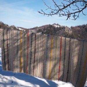 May include: A grey woven blanket with colourful stripes hanging on a wooden fence post in a snowy landscape. The blanket has stripes of yellow, red, green, blue, and purple.