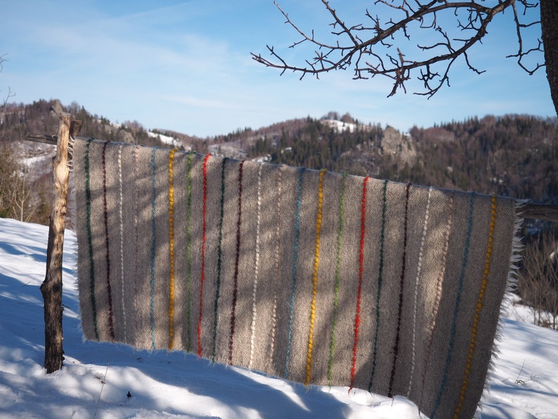 May include: A grey woven blanket with colourful stripes hanging on a wooden fence post in a snowy landscape. The blanket has stripes of yellow, red, green, blue, and purple.