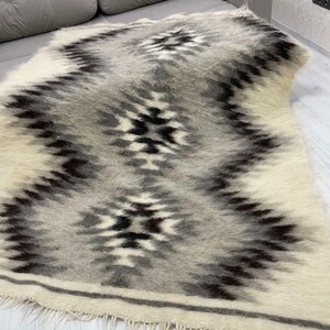 May include: A rectangular, woven rug with a geometric pattern in shades of gray, black, and white. The rug features a central zigzag design with diamond-like shapes. The edges have a fringe detail.