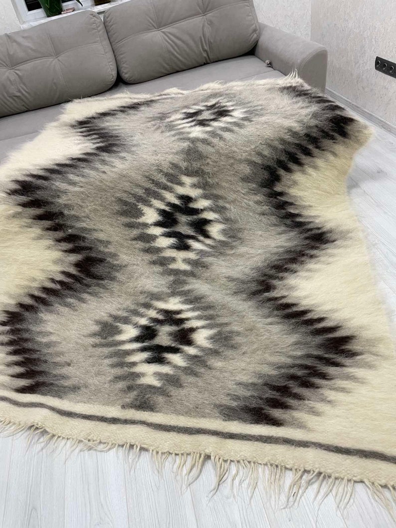 May include: A rectangular, woven rug with a geometric pattern in shades of gray, black, and white. The rug features a central zigzag design with diamond-like shapes. The edges have a fringe detail.