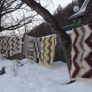 May include: A row of colourful woven blankets hang on a clothesline outside in the snow. The blankets have geometric patterns in shades of brown, white, grey, yellow, and red.