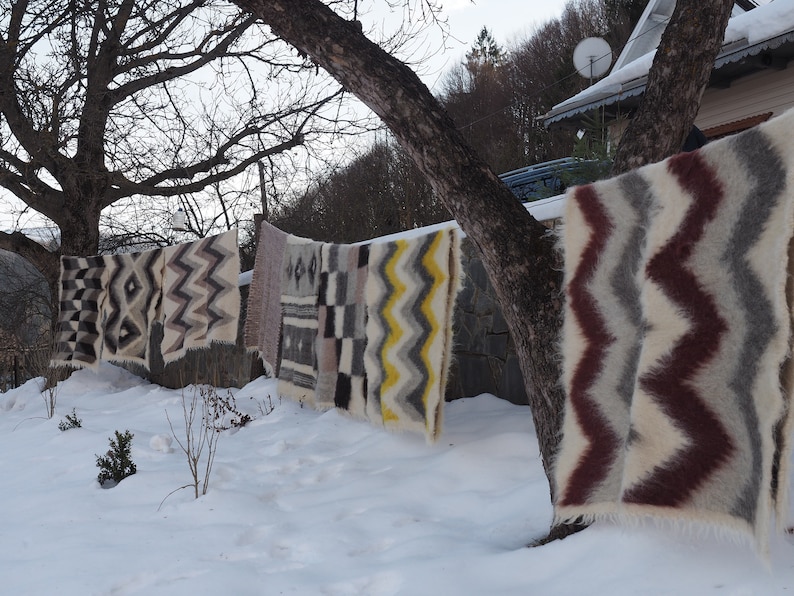 May include: A row of colourful woven blankets hang on a clothesline outside in the snow. The blankets have geometric patterns in shades of brown, white, grey, yellow, and red.