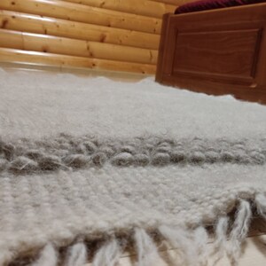 May include: A hand-woven, off-white rug with a thick, textured pile. A darker gray stripe runs across the center, and the rug has a fringed edge. The rug is placed on a wooden floor, near a wooden bed frame.
