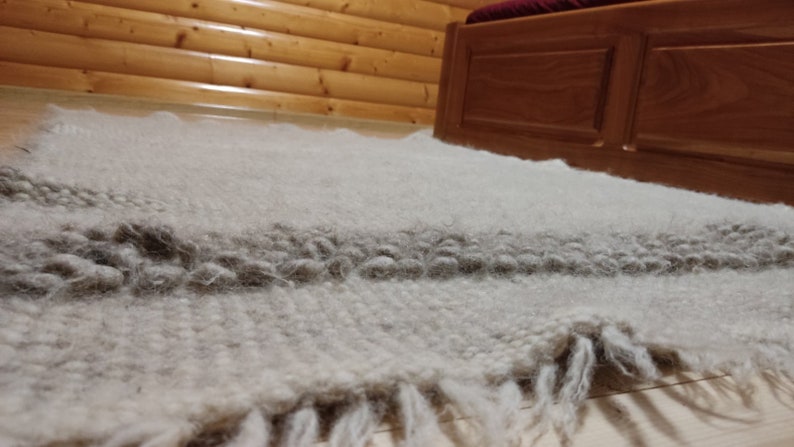 May include: A hand-woven, off-white rug with a thick, textured pile. A darker gray stripe runs across the center, and the rug has a fringed edge. The rug is placed on a wooden floor, near a wooden bed frame.