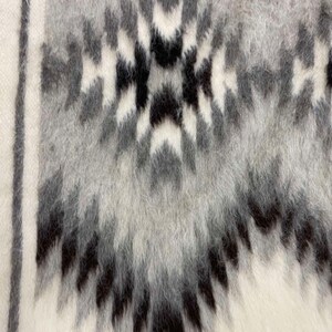 May include: Close-up of a textured textile with a geometric pattern. The design features a repeating motif in shades of gray and black against a white background. The fabric has a soft, fuzzy appearance, suggesting a warm, cozy material.