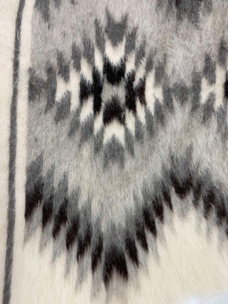 May include: Close-up of a textured textile with a geometric pattern. The design features a repeating motif in shades of gray and black against a white background. The fabric has a soft, fuzzy appearance, suggesting a warm, cozy material.