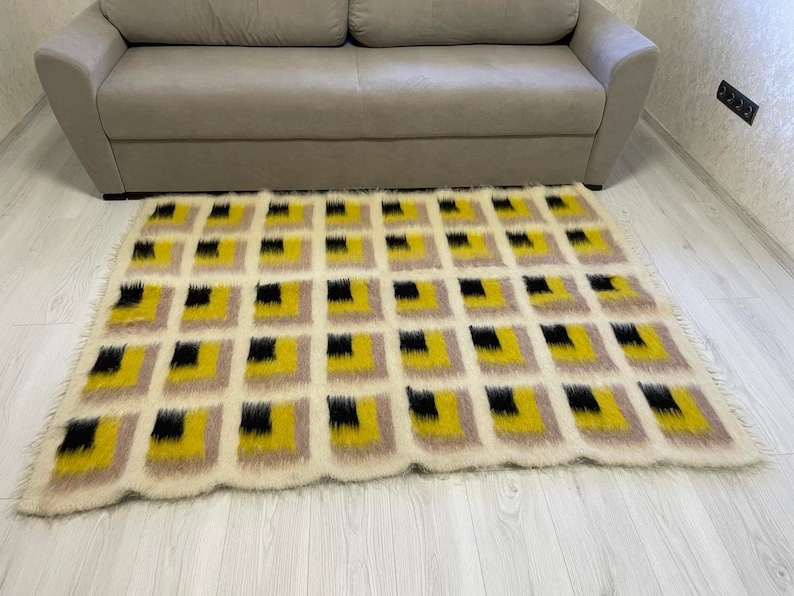 May include: A white, fluffy rug with a geometric pattern of black and yellow squares. The rug is laid out on a light wood floor.