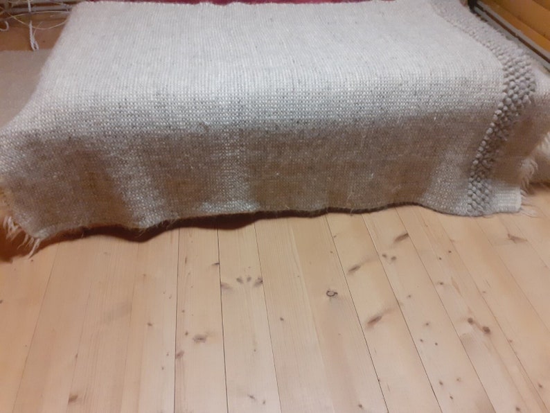 May include: A large, rectangular, off-white woven rug with a textured surface. The rug is draped over an unseen object, with a fringed edge visible. The rug is placed on a light-colored wooden floor.