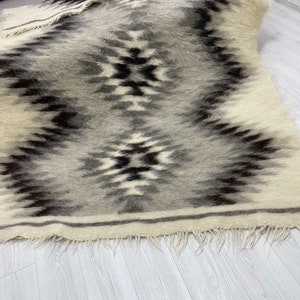 May include: A rectangular, woven rug with a geometric pattern in shades of gray, black, and cream. The rug has a fringed edge and a textured appearance. The design features diamond and zigzag shapes.