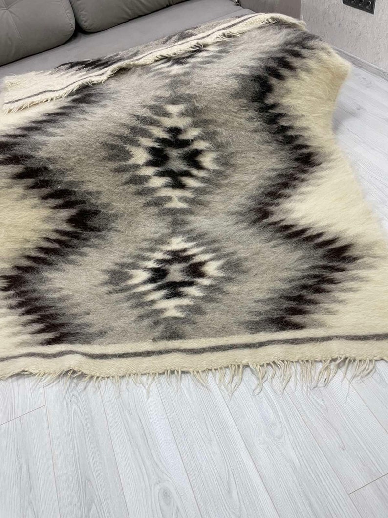 May include: A rectangular, woven rug with a geometric pattern in shades of gray, black, and cream. The rug has a fringed edge and a textured appearance. The design features diamond and zigzag shapes.