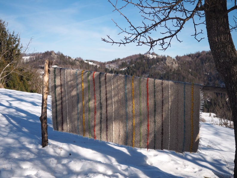 May include: A grey and white striped woven rug with colourful stripes hanging on a wooden post in a snowy landscape. The rug has a rustic, handmade look.